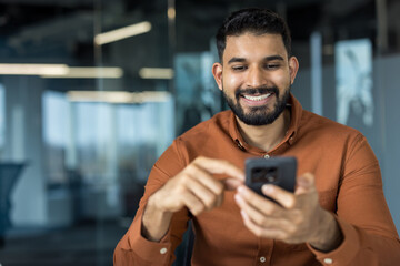 Indian businessman smiling, enjoying connection, holding a mobile phone in a modern office, typing message or checking social media, representing communication and technology