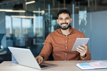 Indian businessman smiles at camera while seated at a desk, working on a laptop and holding a tablet in a modern office, conveying confidence, productivity and digital teamwork