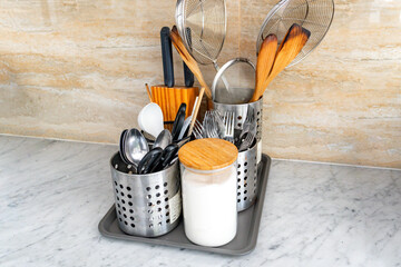 Organized stainless steel kitchen utensil holder on a marble countertop, featuring spoons, knives, wooden tools, and a flour jar