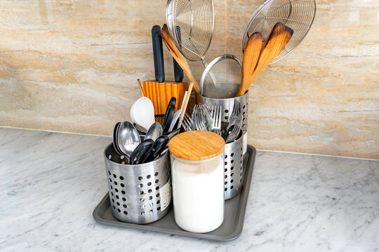 Organized stainless steel kitchen utensil holder on a marble countertop, featuring spoons, knives, wooden tools, and a flour jar - Powered by Adobe