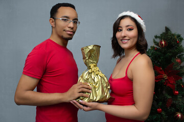 Young adult couple in red outfits exchanging Christmas gifts, smiling, celebrating love, happiness and holiday spirit. Horizontal studio portrait ideal for nativity and xmas romantic present giving.