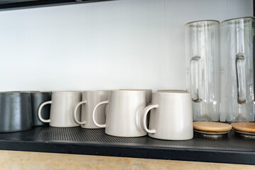 Row of minimalist matte ceramic mugs in gray and dark gray next to clear glass pitchers, neatly arranged on a modern black metal shelf