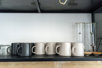 Row of minimalist matte ceramic mugs in gray and dark gray next to clear glass pitchers, neatly arranged on a modern black metal shelf