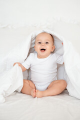 smiling baby girl under a blanket on a white bed, space for text, a small happy child peeking out from under a blanket or going to bed