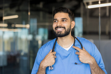 Young indian male medical professional wearing blue scrubs and a stethoscope around his neck,...
