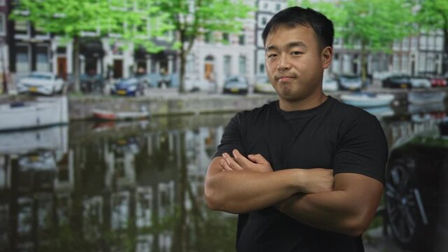 Young man with arms crossed by canal in urban street outdoors near boats and parked cars along tree lined embankment; denial.