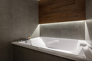 A modern bathroom with a white bathtub set against gray tiled walls and a wooden accent. Soft lighting highlights the sleek design