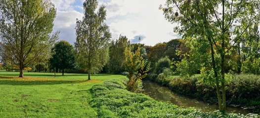 Panoramic landscape photograph of the Rivierenhof in Deurne (Belgium) with grass, green trees, river and sky with clouds.