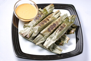 A high-angle view of several grilled otak-otak, wrapped in banana leaves, served on a square plate with a bowl of peanut sauce.