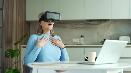 Senior gray haired female plays a game using VR glasses in virtual reality simulator while sitting at the table in kitchen at home. Elderly retired woman uses gestures to control computer programs