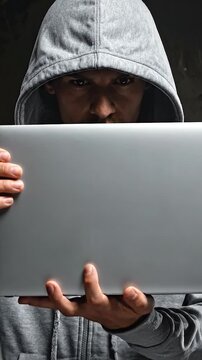 A person wearing a gray hooded sweatshirt holds a laptop, looking intently at the viewer. Dark background, serious expression