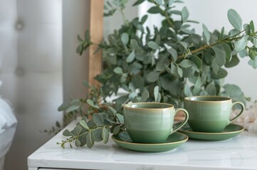 Pair of green mugs with matching saucers and a eucalyptus arrangement on a nightstand. Contemporary home decor.