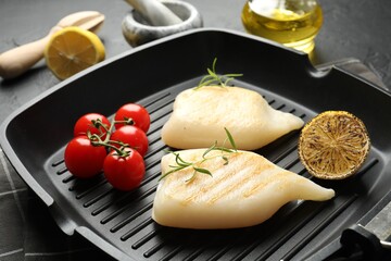 Pieces of tasty grilled squid, tomatoes, lemon and rosemary on dark textured table, closeup