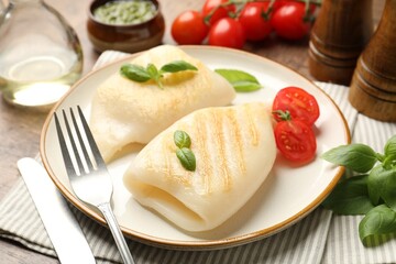Pieces of tasty grilled squid, tomatoes, basil and cutlery on wooden table, closeup