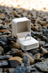 Stunning sapphire engagement ring in a white box placed on rugged dark gravel, symbolizing a bold, outdoor proposal or unique commitment