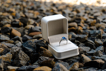 Stunning sapphire engagement ring in a white box placed on rugged dark gravel, symbolizing a bold, outdoor proposal or unique commitment