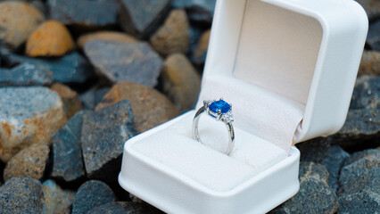 Stunning blue sapphire engagement ring in a white box, set against a backdrop of natural stones. Perfect for luxury and romance themes