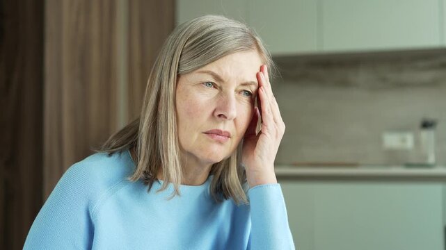 Close up senior gray haired female suffering from headache sitting at kitchen table at home. Elderly retired woman massaging her temples with hands. She is tired, has high blood pressure, feels pain