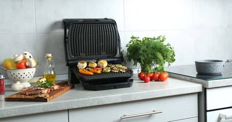 Electric grill with delicious vegetables and steaks on light table in kitchen