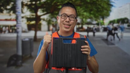 Man in reflective orange vest holding black case and showing thumbs up on street; confidence service.