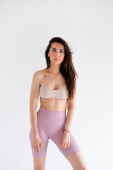Obraz premium Sports woman standing in activewear with abs muscles, looking at camera isolated on white background. Caucasian 30s fitness girl wears beige top and pink leggings. Slim fit brunette with blue eyes.