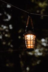 A glowing vintage lantern hanging by a rope in the quiet forest twilight. Captured with a shallow depth of field, the warm light contrasts beautifully against the dark, moody background of trees