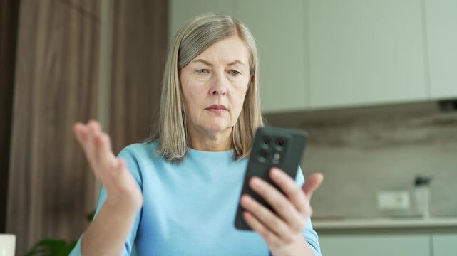 Close up of confused senior female with gray hair having difficulty using smartphone sitting in kitchen at home. Worried puzzled elderly woman has a problem complexity working on application on phone