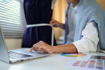 A stylishly dressed male designer creates a new collection of clothes, measures the parameters of clothes with a measuring tape and writes them down on laptop while standing in his studio.