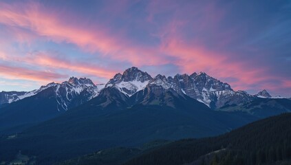 Snow-capped summits towering over dense green woods beneath a colorful sky in a stunning mountain landscape.