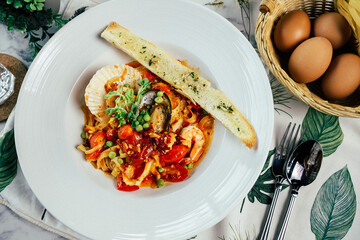 A plate of delicious seafood pasta with vegetables and a slice of garlic bread, served with a basket of eggs. This image has an overhead view and a vibrant color palette.