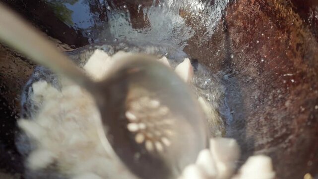 In slow motion, a slotted spoon stirs melting lamb tail fat in a hot cauldron outdoors during the first stage of traditional pilaf preparation, as the fat renders and bubbles in the heat