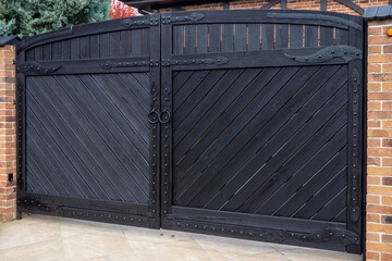 Large black metal driveway gate with diagonal pattern and brick pillars, private property entrance. For architecture, fencing systems, property development, urban design, security concepts. Real photo