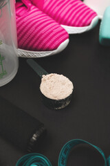 Scoop of whey protein powder, teal dumbbells, pink sneakers, jump rope, shaker bottle and measuring tape on dark background with copy space, fitness equipment and sports nutrition concept