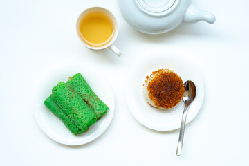 A top-down view of Indonesian snacks, Kue Dadar Gulung and Ketan Serundeng, on white plates with a cup of tea and a teapot on a white background.
