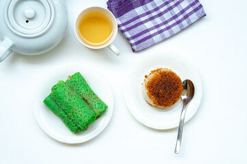 A top-down view of Indonesian snacks, Kue Dadar Gulung and Ketan Serundeng, with a cup of tea and a teapot on a white background.