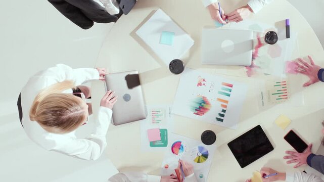 High perspective of business meeting with charts, laptops, and sticky notes on round table. Concept of teamwork, data-driven management, idea generation, and analytical collaboration.