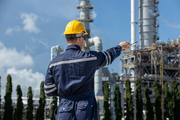 Back view of engineer in safety uniform pointing at refinery plant while using walkie-talkie representing industrial field control, communication, supervision safety management and oil gas operations.