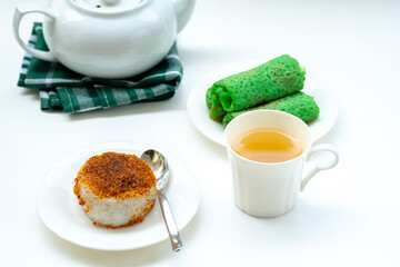 Indonesian snacks, Ketan Serundeng and Kue Dadar Gulung, are served on white plates with a cup of tea and a teapot, all on a white background.