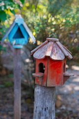 Weathered red birdhouse on wooden post in garden, with blurred blue birdhouse in background. Rustic charm and nature setting