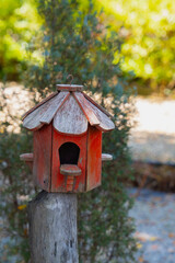 Rustic red birdhouse with weathered appearance stands on wooden post in garden setting. background features blurred greenery and sunlight