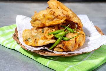 A pile of Indonesian fried vegetable fritters, bakwan, is served on a small wicker plate with paper and green chilies, sitting on a green checkered cloth.