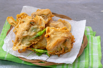 A small stack of Indonesian tahu isi (deep-fried stuffed tofu) with a crispy batter coating and green chili peppers on a woven bamboo plate.