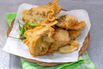 A small stack of Indonesian tahu isi (deep-fried stuffed tofu) with a crispy batter coating and green chili peppers on a woven bamboo plate.