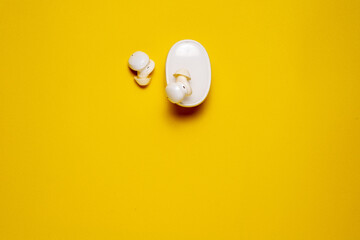 A top-down view of white wireless earbuds, one resting on the closed charging case and the other beside it, on a bright yellow background.