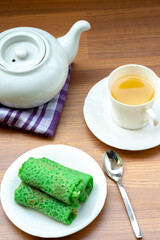 A plate with three green dadar gulung, Indonesian rolled crepes, served with a cup of tea and a white teapot on a wooden table.