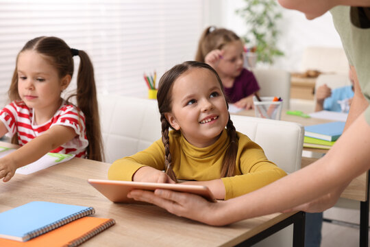 Teacher and smiling little girl with tablet at wooden desk in elementary school, closeup