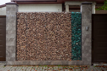 Gabion wall with brown and green stones between tiled pillars near garage entrance. Design fences, fencing of the homestead plot. Real photo