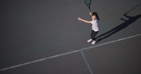 A young girl with focused intensity plays tennis, her dynamic pose captured as she reaches for the ball on an outdoor court under clear skies.