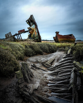 Decaying wooden shipwreck and rusting metal hull on marshes under overcast sky