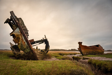 Decaying wooden shipwreck and rusting metal hull on marshland under overcast sky
